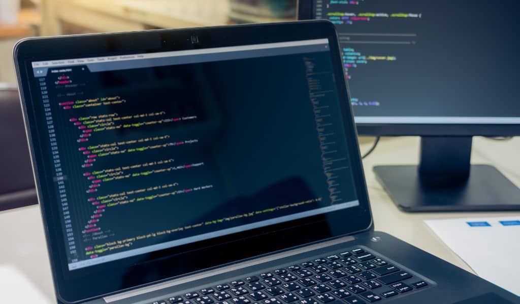 parallelstaff- A laptop displays code in a text editor with colorful syntax highlighting, while a second monitor in the background shows more lines of code—an efficient workspace that suggests nearshore software development or web programming.