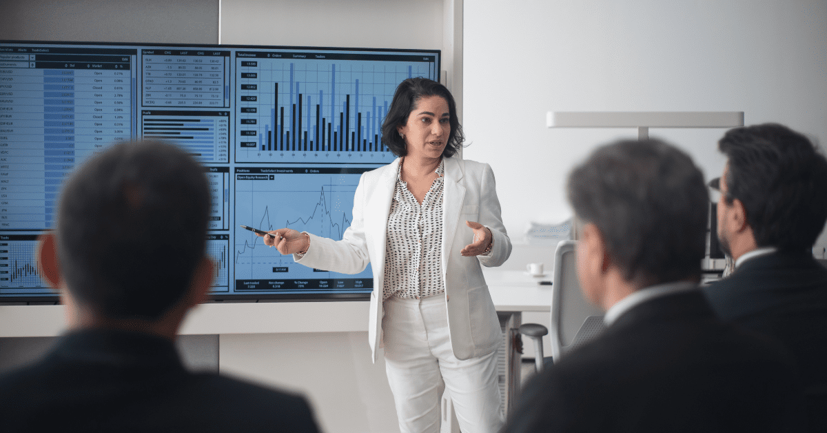 A businesswoman in a white suit presenting software budget analysis and performance charts to a group of executives.
