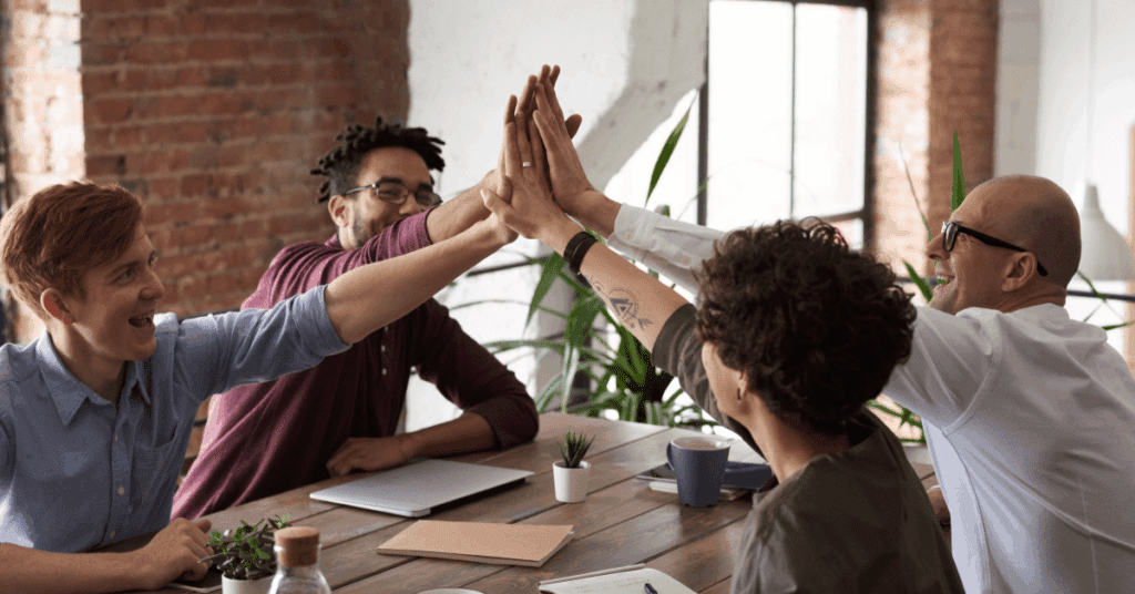 Group of diverse professionals high-fiving around a work table, reflecting strong team synergy with a trusted outsourcing partner.