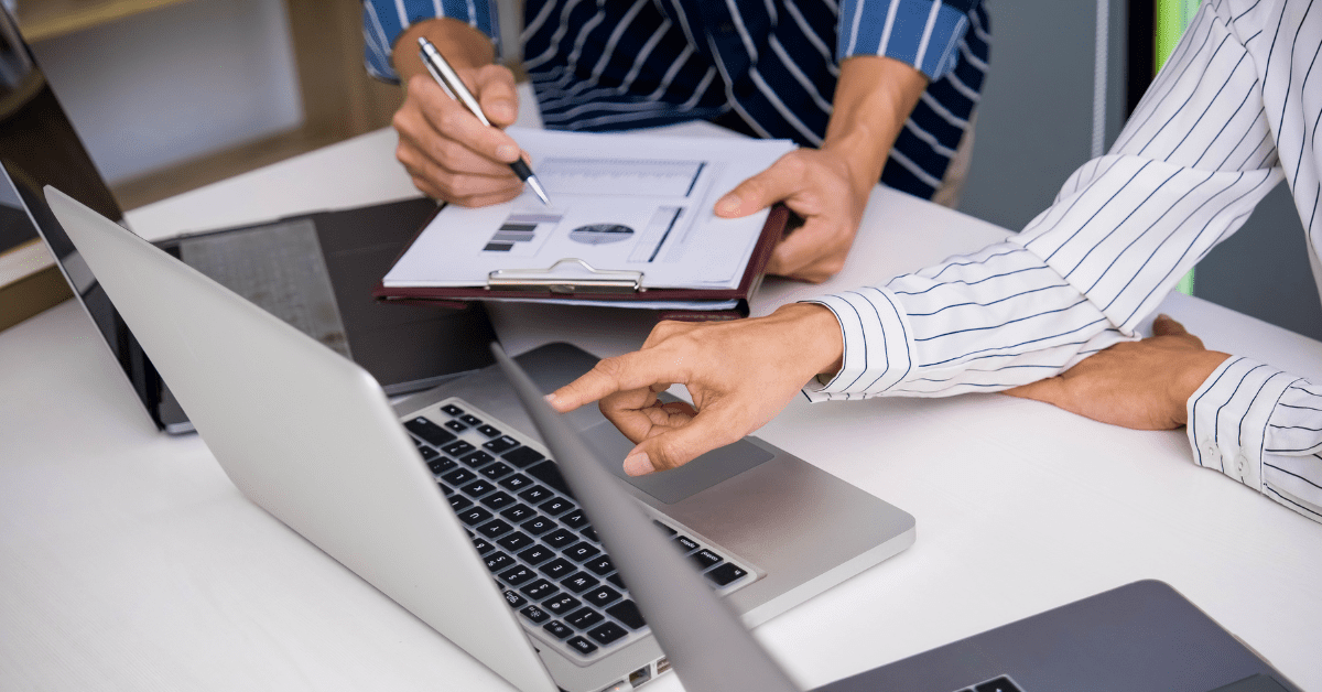 Close-up of two professionals analyzing charts and data on a clipboard and laptop, discussing efficient IT budget strategies.