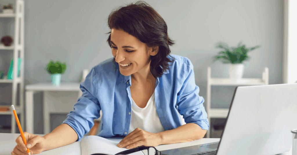 Woman smiling and writing notes while working remotely with a laptop on desk.