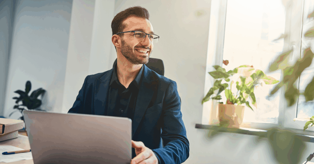 Confident man smiling while working on his laptop in a bright office with plants.