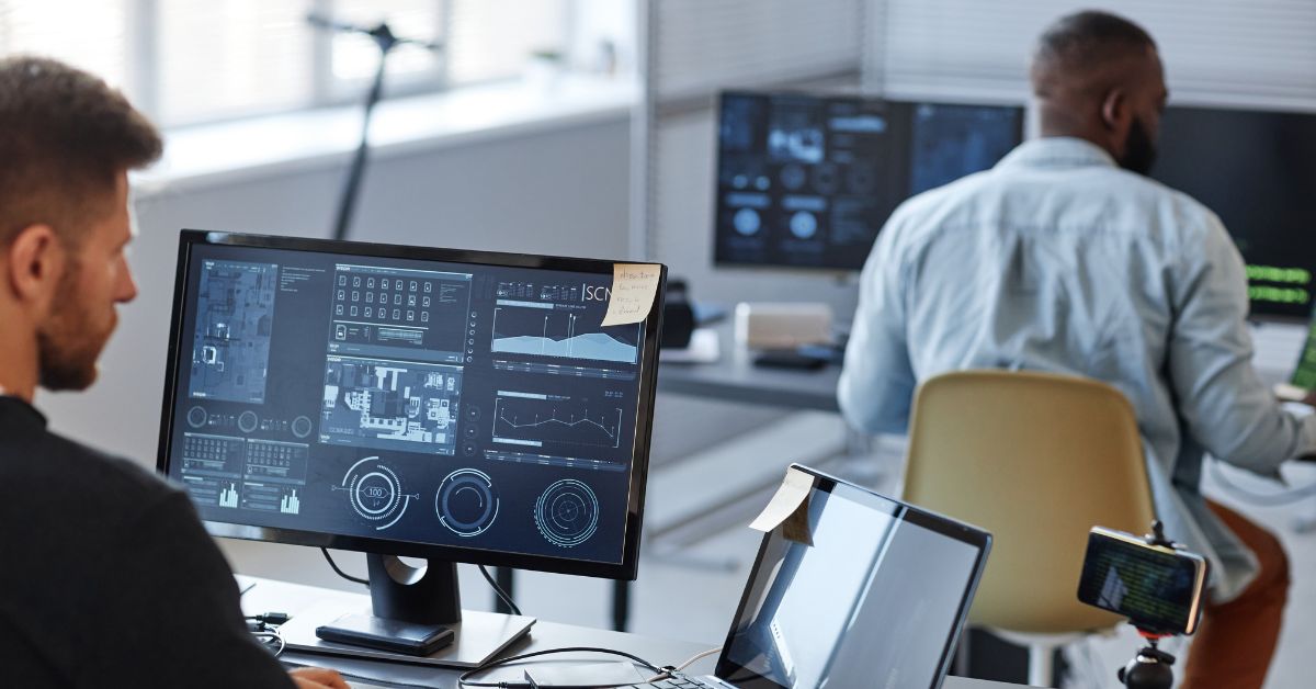 Two AI engineers working in a modern office with data dashboards and technical analytics on their screens.