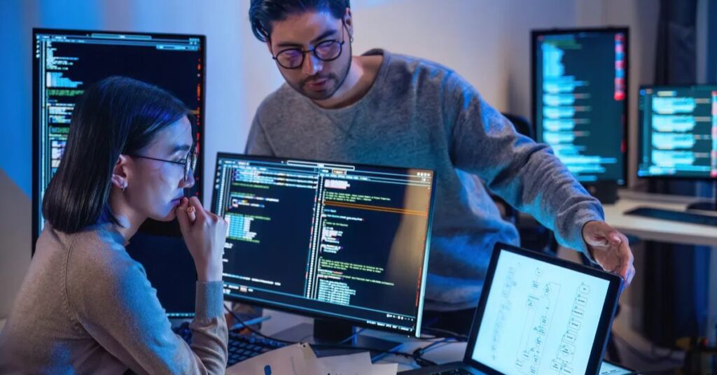 Two AI developers collaborating on code and project planning in a multi-monitor setup.