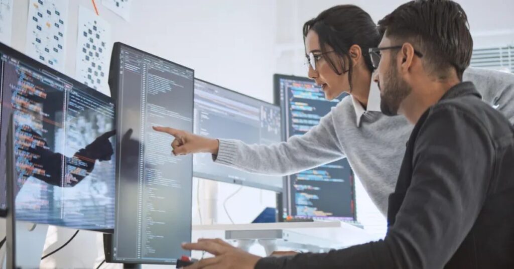 Two developers discussing and analyzing AI code on large screens in a collaborative environment.