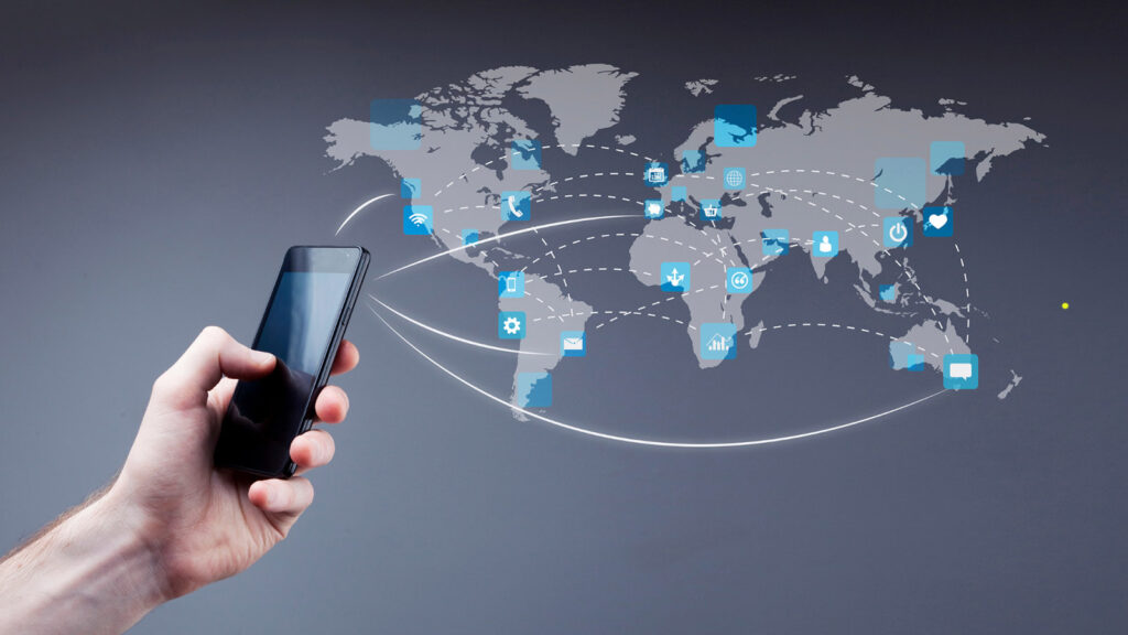 parallelstaff- A hand holds a smartphone in front of a world map with digital icons and curved lines, symbolizing global connectivity, communication, and data exchange—key elements for a remote staffing agency leveraging mobile technology.