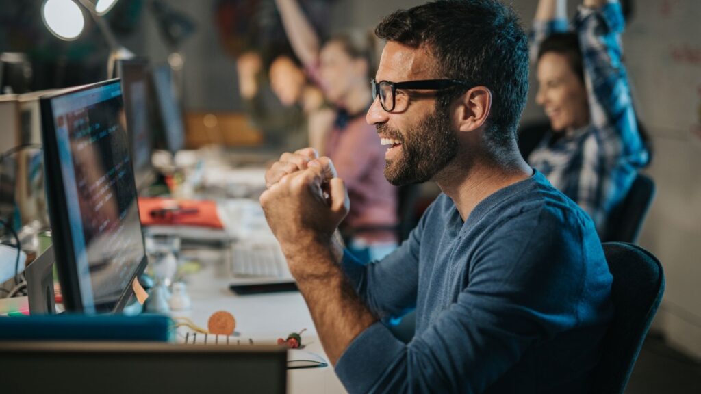 parallelstaff- A man wearing glasses and a blue shirt smiles and clenches his fists in excitement while looking at a computer screen, celebrating recruitment strategies to attract and retain talent with colleagues in an office setting.