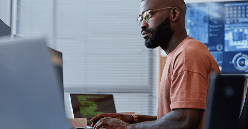 parallelstaff- A man wearing glasses and a brown t-shirt works at a computer in an office, with code on his laptop screen—capturing the daily life of a software developer vs software engineer—while digital interface graphics appear on a monitor behind him.