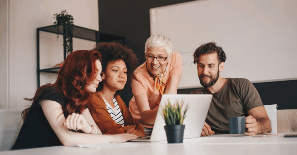 parallelstaff- Four people sit together at a table, smiling and looking at a laptop screen—perhaps comparing software developer vs software engineer roles. A plant and coffee mug are on the table, with shelves and a whiteboard in the background.
