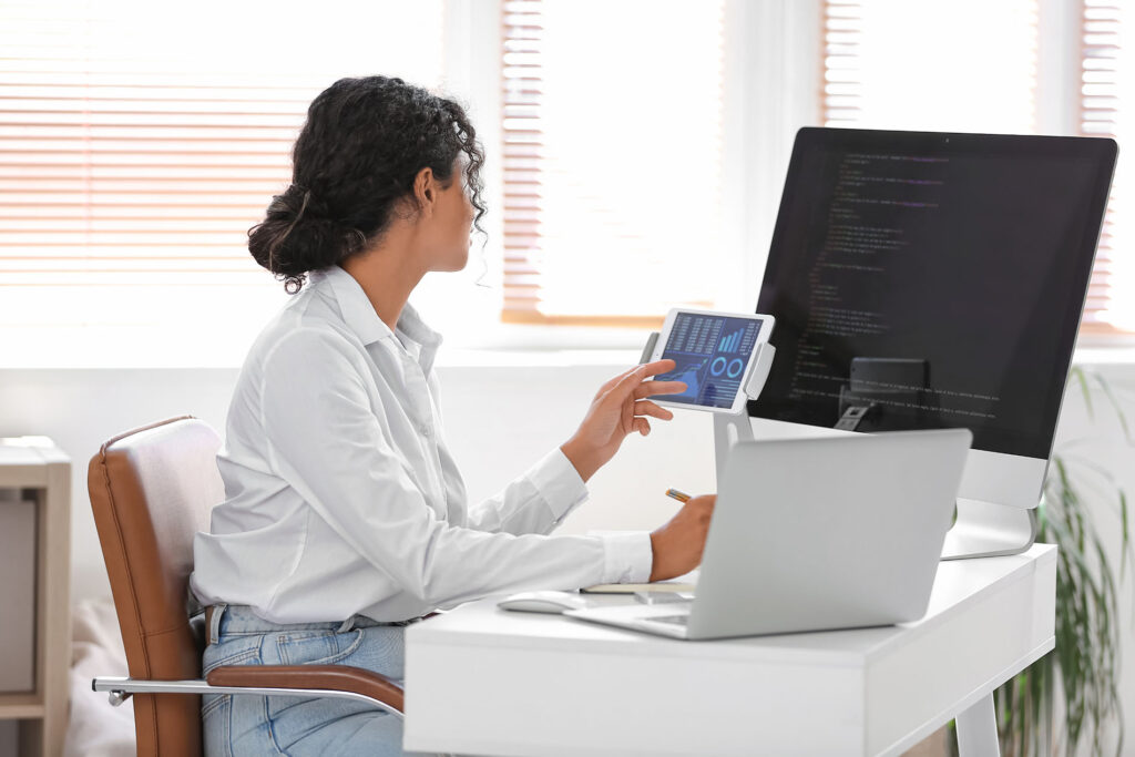 parallelstaff- A woman sits at a desk with a computer, tablet, and laptop, analyzing data and code on her screens—an example of what a data engineer does in a bright office with large windows.
