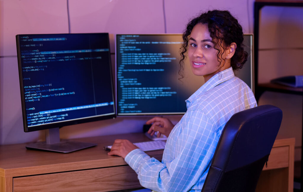 parallelstaff- A woman sits at a desk, smiling at the camera, with two computer monitors displaying lines of code—capturing the essence of what a data engineer does—in a modern, softly lit workspace. She wears a light blue, plaid shirt.
