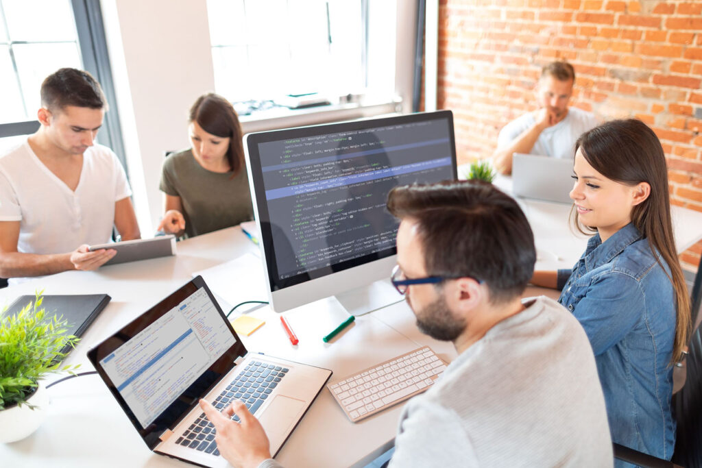 parallelstaff- Five people work together in a modern office. Two focus on a computer displaying code, while others use laptops and tablets around the table—a bright, collaborative space ideal to hire a dedicated development team.