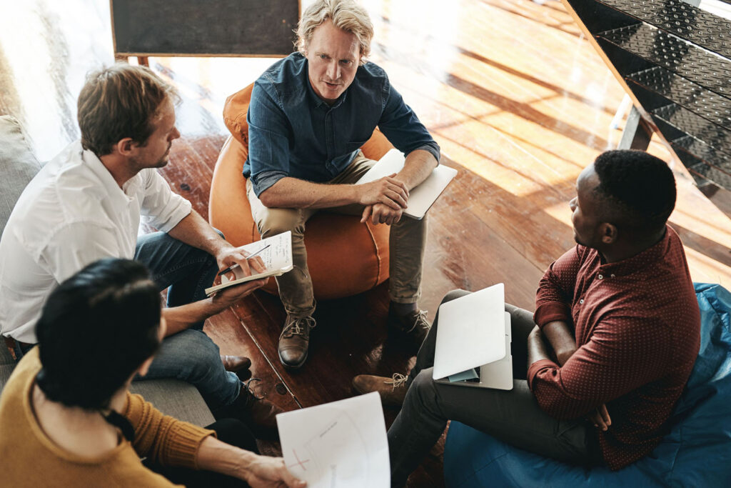 parallelstaff- Four people sit in a casual circle on bean bags, engaged in discussion. Holding notebooks and a tablet, they brainstorm ideas to hire a dedicated development team in their relaxed, modern office setting.