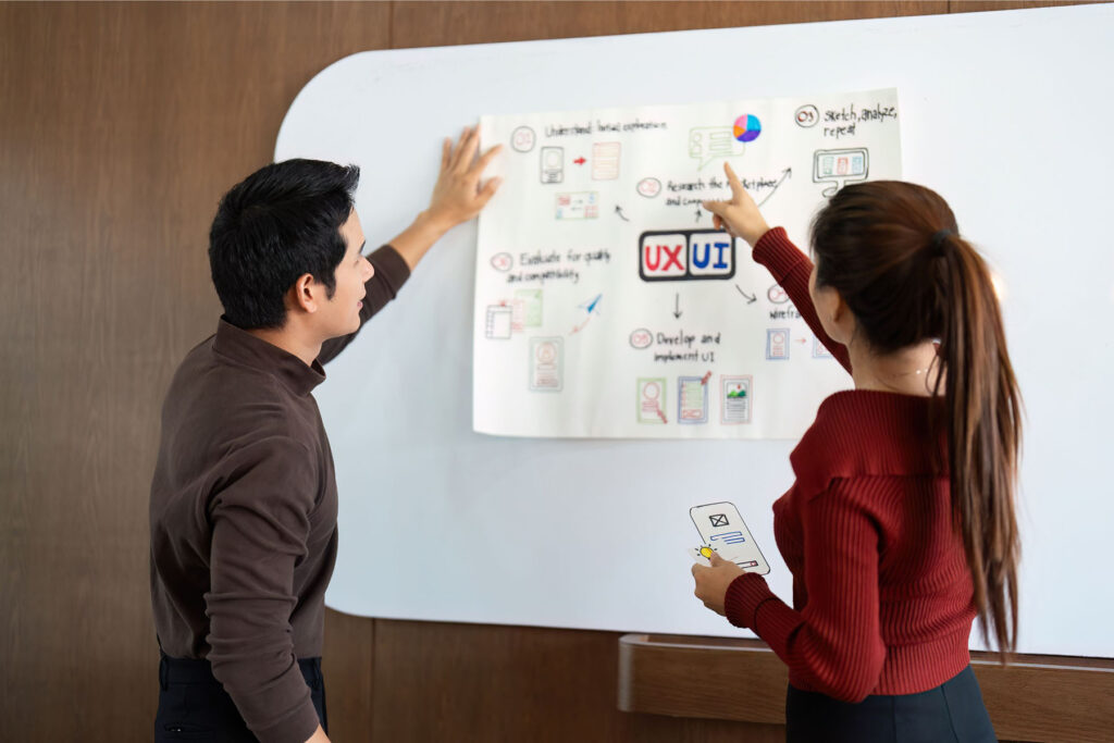 parallelstaff- Two people stand by a whiteboard, discussing a UX/UI design flowchart. They examine visual elements and steps outlined on the paper, illustrating insights from the "What Does a UI/UX Designer Do? Hiring Guide." One points to the chart while the other holds a phone mockup.