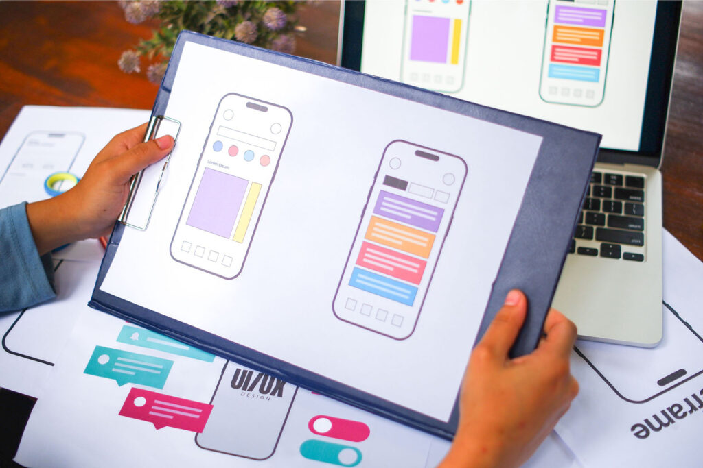 parallelstaff- A person holds a clipboard with smartphone app wireframes, reviewing colorful mockup designs—illustrating What Does a UI/UX Designer Do? Hiring Guide materials; a laptop and more design sketches with interface elements are on the desk.