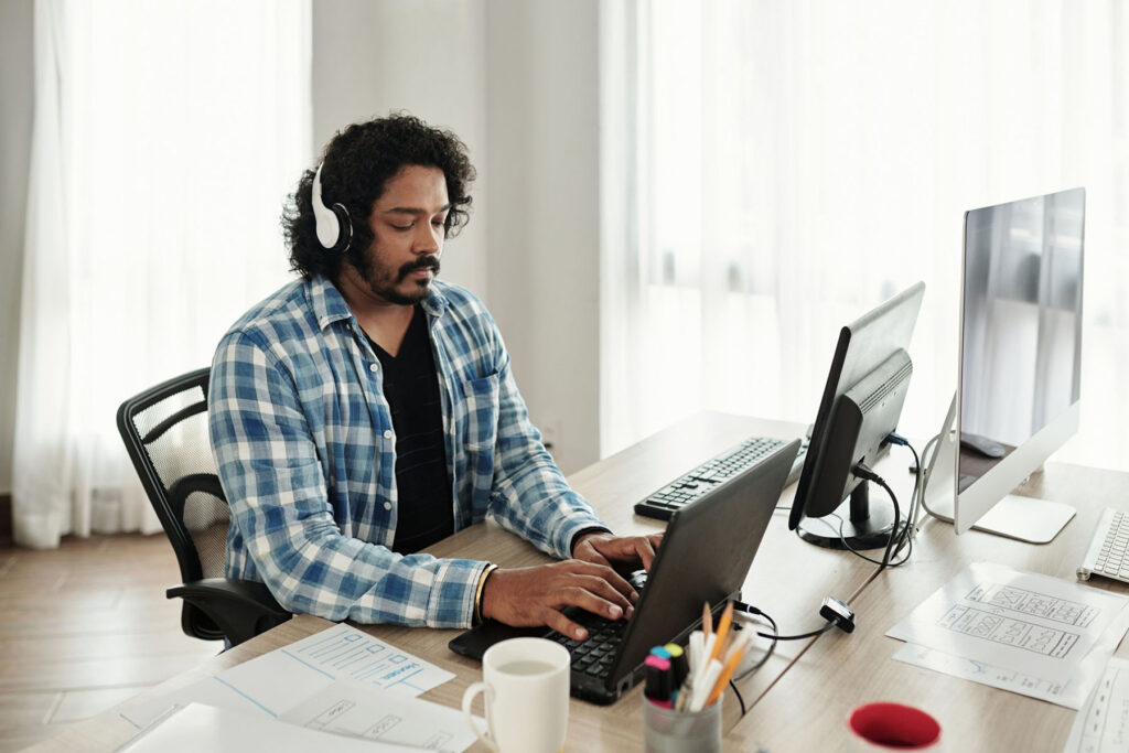 parallelstaff- Man wearing headphones and a blue plaid shirt works on a laptop at a desk with two monitors, papers, and office supplies in a bright, modern office—researching how to hire a front end developer efficiently.