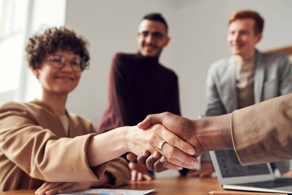 parallelstaff- Two people are shaking hands at a meeting table while two colleagues look on and smile, highlighting a successful business agreement—perfect for teams looking to hire iOS developers for their next project.