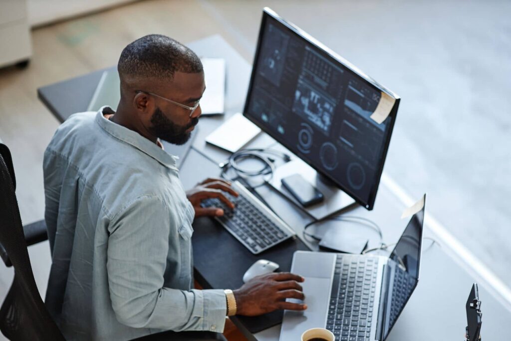 parallelstaff- A man wearing glasses works at a desk with two monitors, a laptop, and various electronic devices. Focused on charts and data, he uses both the keyboard and mouse—an ideal scene for teams looking to hire iOS developers.