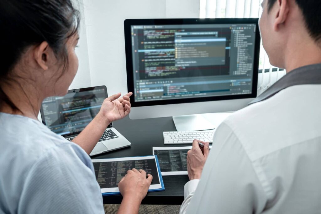 parallelstaff- Two people sit at a desk working together on computer code displayed on a large monitor, typical of an Offshore Software Development team. One gestures toward the screen while the other uses the keyboard, with a laptop and tablet also on the desk.