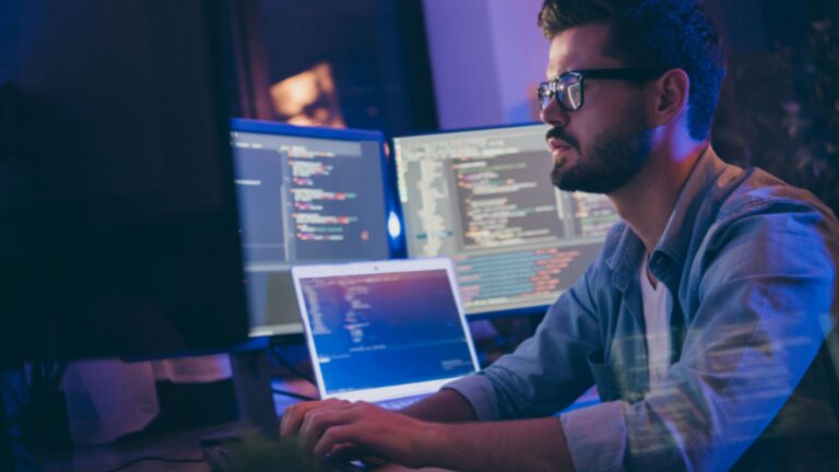 parallelstaff- A man wearing glasses is typing on a keyboard, sitting at a desk with multiple monitors displaying lines of code in a dimly lit room, suggesting he is working on computer programming or software development.
