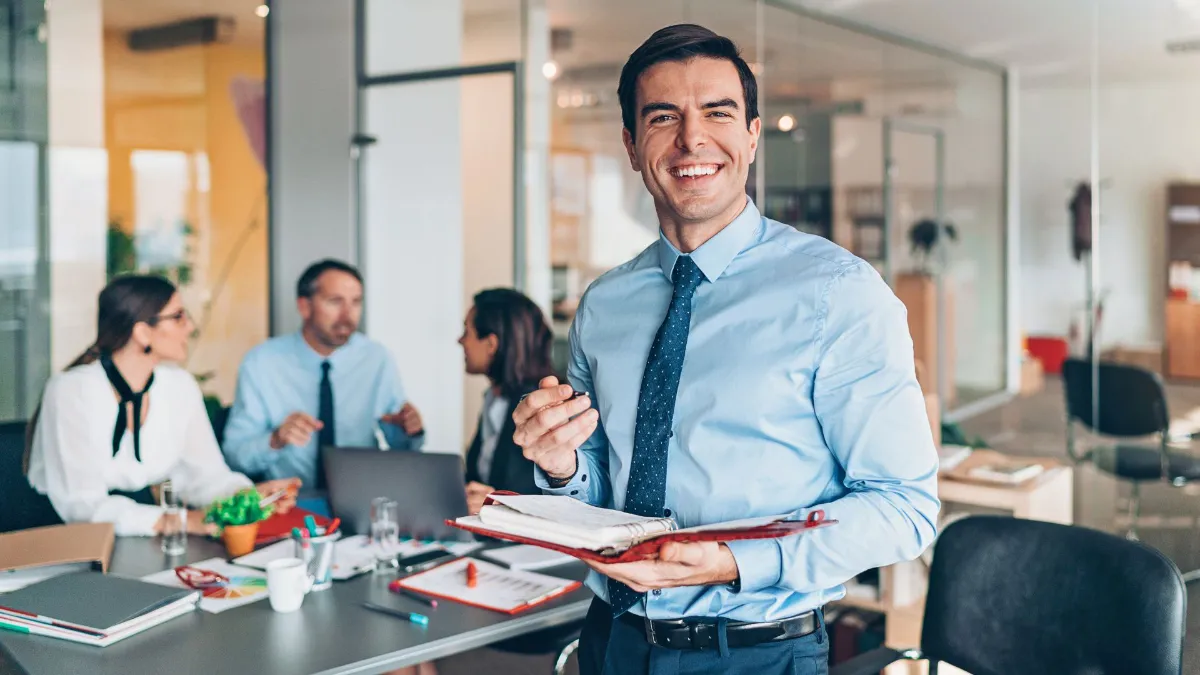 parallelstaff- A smiling man in business attire holds an open notebook in a modern office, while three colleagues sit at a table in the background having a discussion.