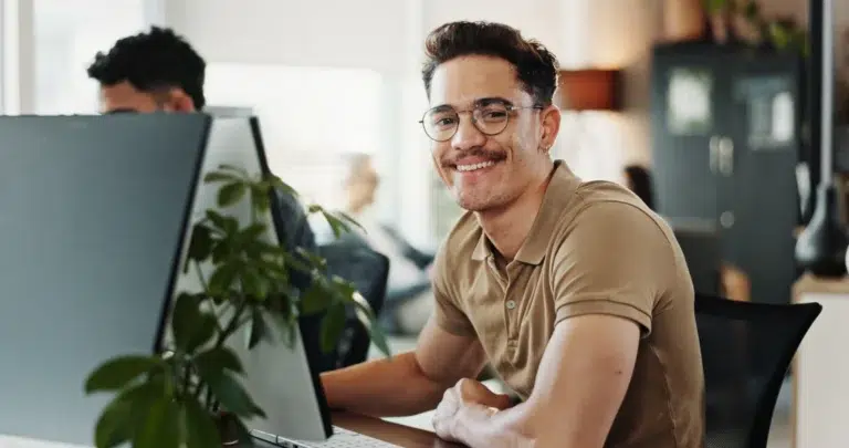 parallelstaff- A smiling man wearing glasses and a brown polo shirt sits at a desk with a computer, looking toward the camera. A green plant is beside the monitor, and an office environment is visible in the background.