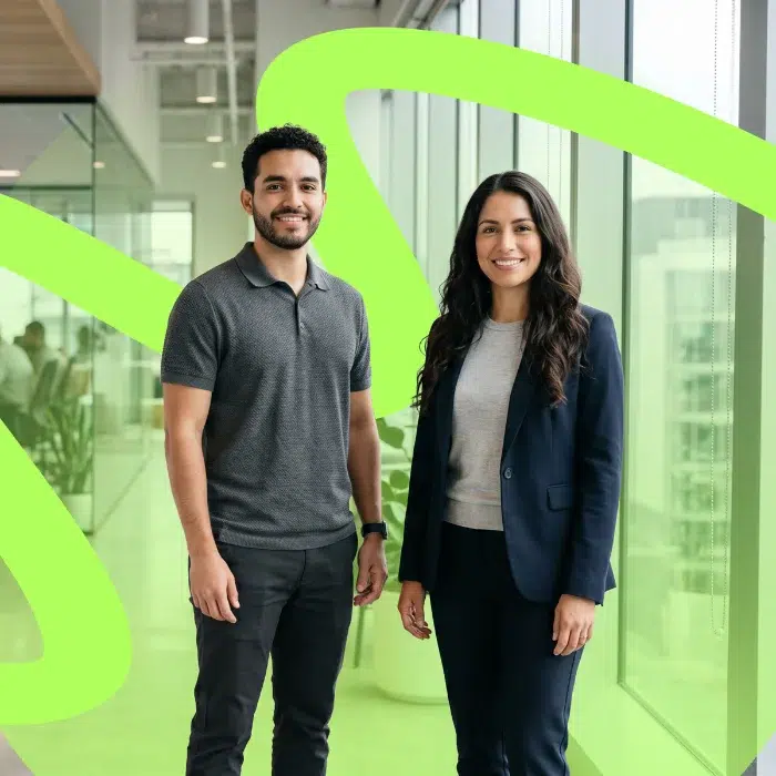 parallelstaff- Two people, a man in a gray polo and black pants and a woman in a navy suit and gray top, stand smiling in a modern office hallway with large windows and green abstract lines overlaid.