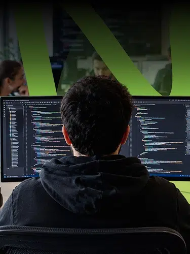 parallelstaff- A person with dark hair wearing a black hoodie sits at a desk in front of two large monitors displaying colorful code, with a green abstract shape in the background.