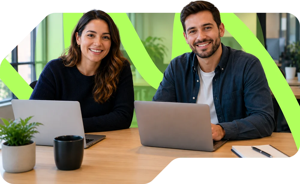 parallelstaff- Two people sit at a desk with laptops, smiling at the camera. A potted plant, coffee mug, and notepad are on the table. The background is bright and modern with green accents.