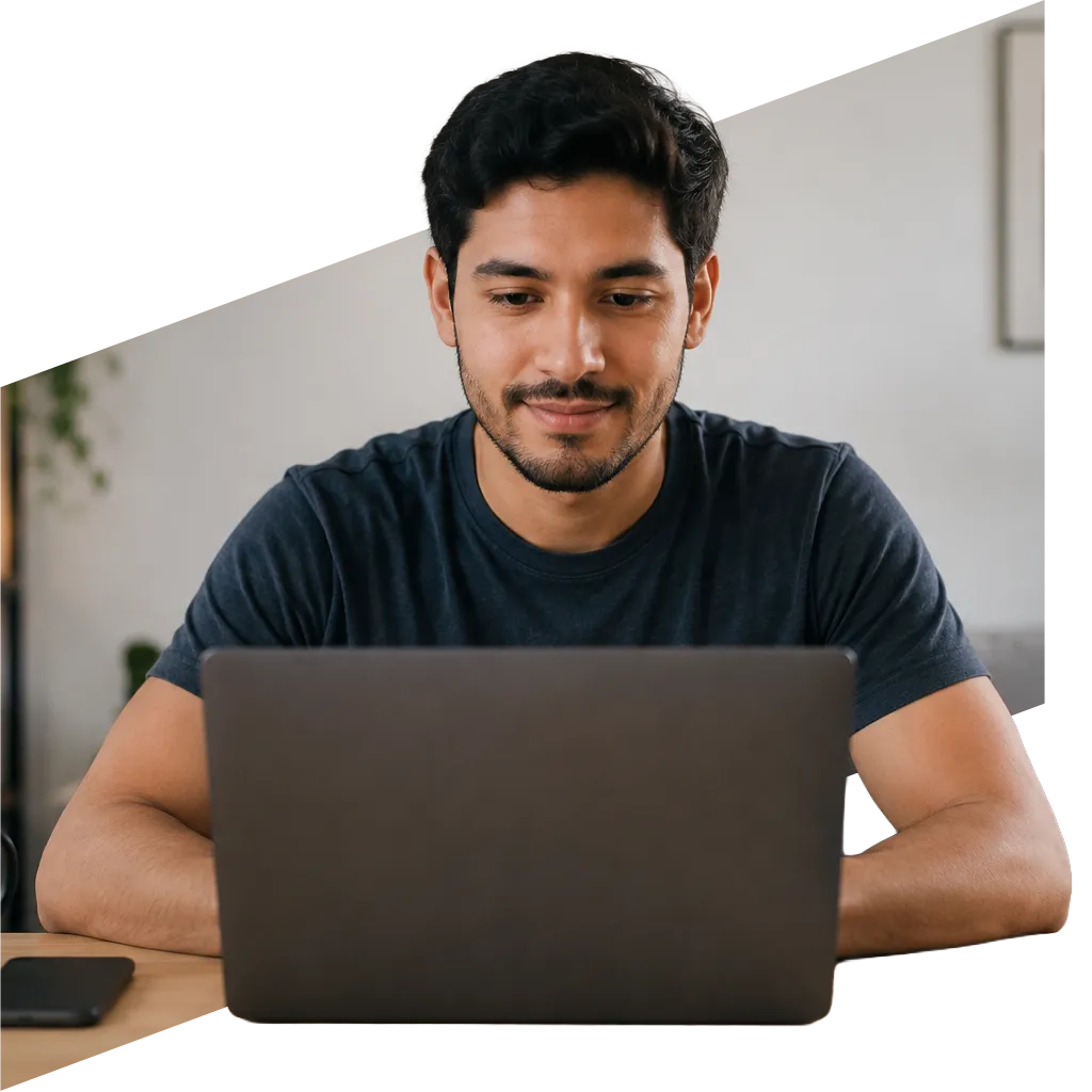 parallelstaff- A man with dark hair and a beard sits at a desk, smiling while looking at a laptop screen. The background is softly blurred, suggesting a home or office environment.