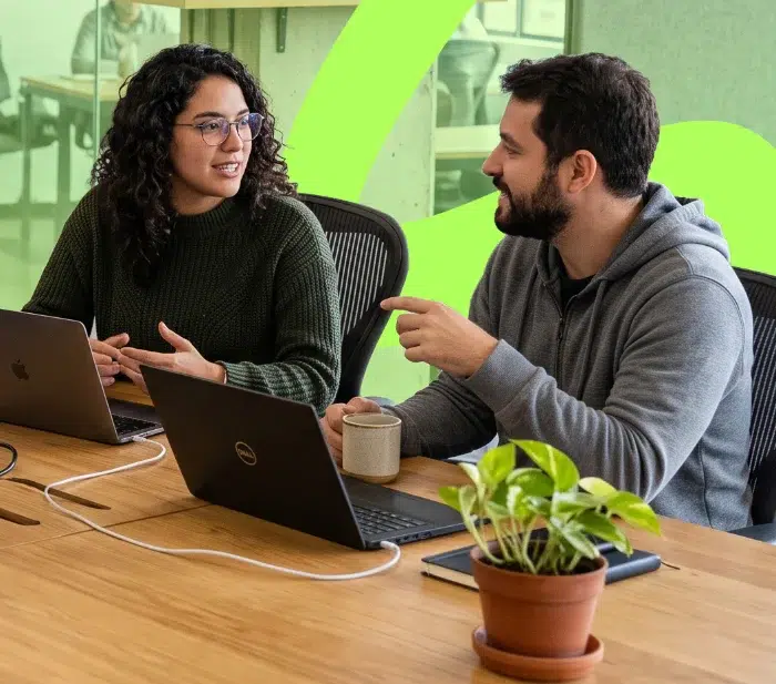parallelstaff- Two people sit at a table with laptops, talking and smiling. One holds a mug. A potted plant and a phone are on the table. Bright green shapes decorate the wall in the background.