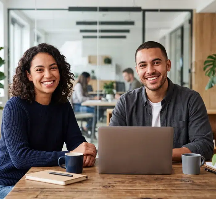parallelstaff- Two people sit at a wooden table with a laptop, notebook, and coffee mugs, smiling at the camera in a modern office setting with glass walls and plants.