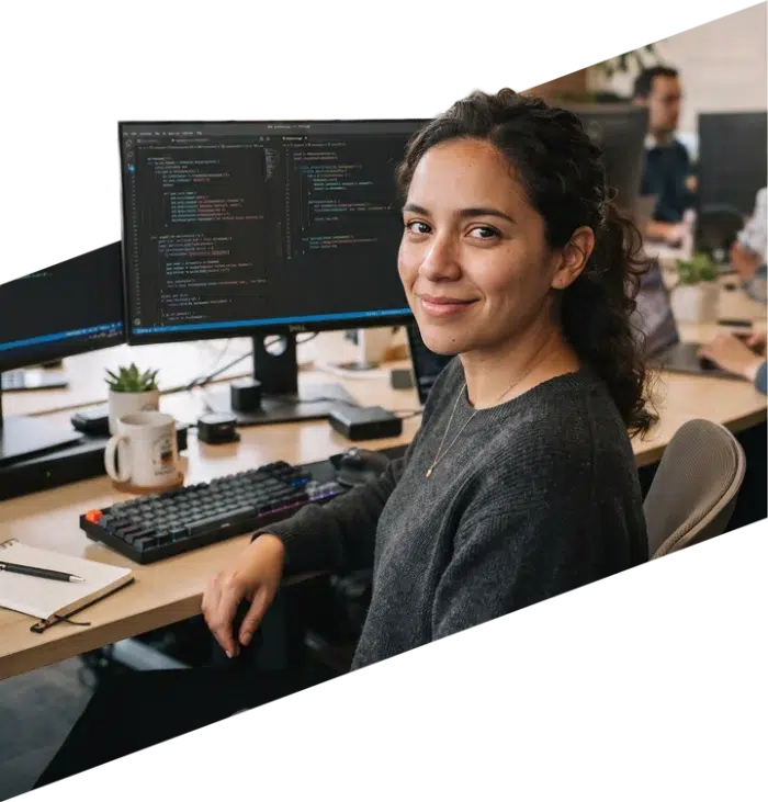 parallelstaff- A woman with curly hair sits at a desk with two monitors displaying code, smiling at the camera. A keyboard, mug, plant, and notebook are on the desk. Other people work in the background.