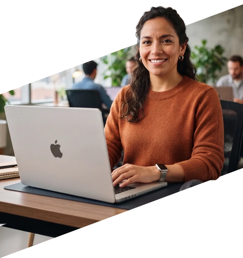 parallelstaff- A woman in an orange sweater smiles while working on a laptop at a desk in a modern office. Other people are visible working in the background.